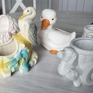 May include: Four ceramic planter pots shaped like a duck, a stork, a baby carriage, and a dog pulling a wagon. The pots are all white with painted details. The duck and dog planters have a light gray glaze.
