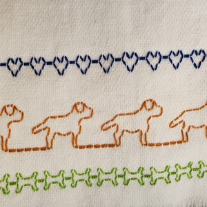 Pode incluir: Tecido branco com desenhos bordados. Apresenta fileiras de corações azuis, silhuetas de cães castanhos e formas de ossos verdes. A costura cria um padrão decorativo texturizado.