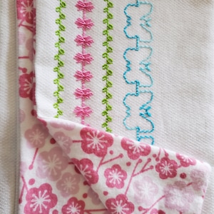 Può includere: Un asciugamano da cucina bianco con ricami verdi, rosa e blu. L'asciugamano è piegato su un tessuto rosa e bianco con un motivo floreale.