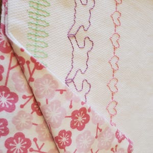 Puede incluir: Una tela blanca con diseños de bordado rosa y verde. La tela está doblada sobre una tela blanca con un patrón floral rosa.