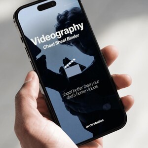 May include: A smartphone displaying a digital cover for a Videography Cheat Sheet Binder. The screen features white text reading Videography Cheat Sheet Binder, a clapperboard icon, and the text shoot better than your dad's home videos, with avco studios at the bottom.