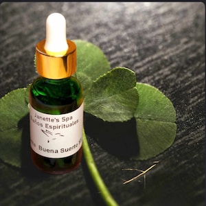 May include: A small green glass bottle with a gold lid and a white label that reads "Janette's Spa Baños Espirituales Buena Suerte". The bottle is sitting on a dark surface with a green four-leaf clover.