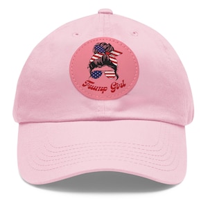 May include: Pink baseball cap with a round patch featuring a woman with a bun in her hair, wearing sunglasses and a red, white and blue headband. The patch also has the text "Trump Girl" in red and white.