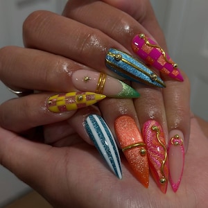 May include: Stiletto-shaped nails featuring a variety of designs including pink and yellow checkered patterns, blue and white stripes, glittery orange, and solid nude bases. The nails are decorated with gold metallic lines, small gold spheres, and star accents.