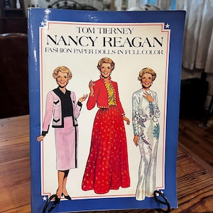 May include: A book titled "Nancy Reagan Fashion Paper Dolls in Full Color" by Tom Tierney. The cover features illustrations of Nancy Reagan in various outfits, set against a blue background with white stars.
