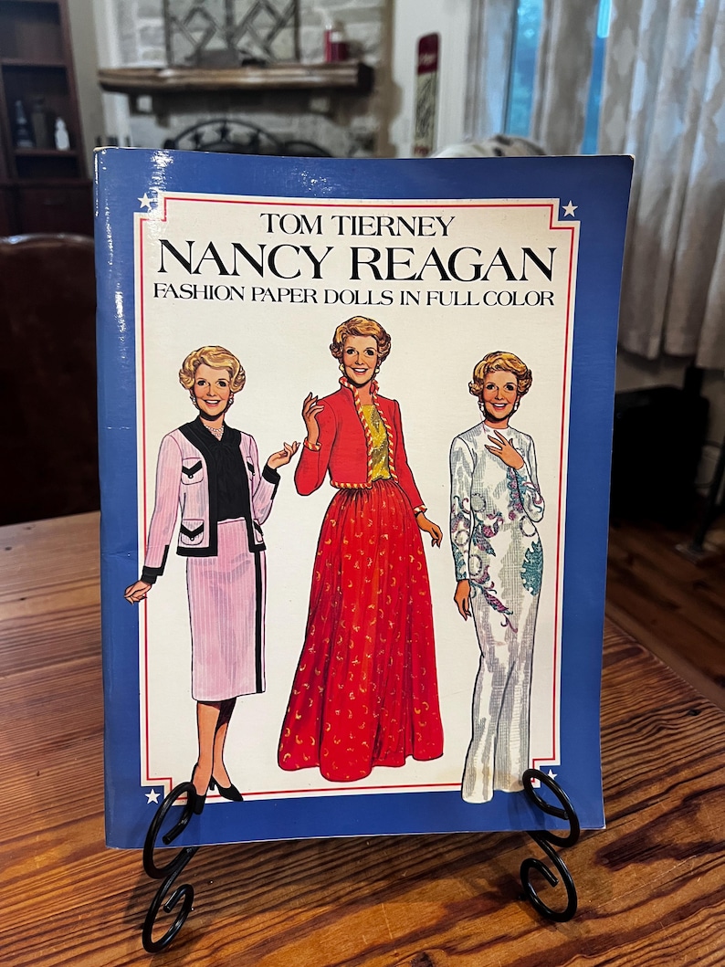 May include: A book titled "Nancy Reagan Fashion Paper Dolls in Full Color" by Tom Tierney. The cover features illustrations of Nancy Reagan in various outfits, set against a blue background with white stars.