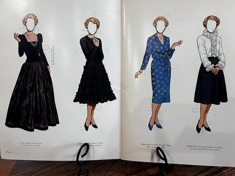 May include: Vintage paper dolls featuring four different outfits. The outfits include a black evening gown, a black dress with a tiered skirt, a blue two-piece dress with a floral pattern, and a white blouse with a black A-line skirt.