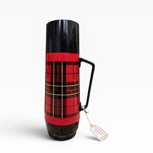 Op de afbeelding: Een vintage thermoskan met een zwarte handgreep en deksel. De behuizing heeft een rood-zwart ruitpatroon met gele accenten. Een klein label is aan de handgreep bevestigd.