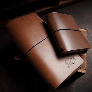 May include: Two brown leather journals with black elastic bands. The larger journal features a stamped graphic of a turtle carrying a mushroom on its shell with the text "MAKESUPPLY U.S.A." The covers are smooth, matte, and rectangular.