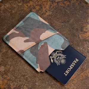 May include: A camouflage patterned passport holder with visible white stitching along the edges. The holder is partially open, revealing a dark blue passport with a gold emblem and the word Passport printed on the front cover.