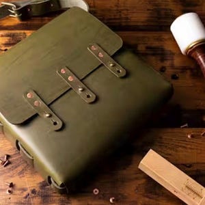 May include: An olive green leather satchel with a flap closure secured by three vertical leather straps and copper rivets. The bag features an adjustable shoulder strap and is accompanied by a white mallet and a wooden tool on the surface.
