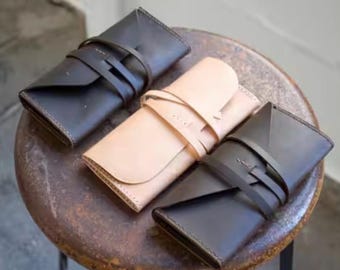 May include: Three rectangular leather cases with fold-over flaps and wrap-around leather cord closures. Two cases are dark brown and one case is light tan. Each case features visible stitching along the edges and a smooth, matte leather surface.
