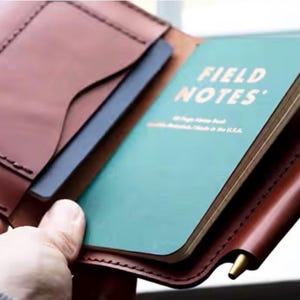 May include: A brown leather notebook cover featuring a teal Field Notes memo book with white text. The cover includes a side slot for a gold-toned metal pen and an interior pocket for cards or documents, showcasing visible stitching along the edges.