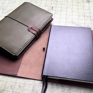 May include: Two leather notebook covers are displayed. One is a closed olive green cover with a dark brown strap closure. The other is an open dark brown cover holding a dark purple notebook with a black ribbon bookmark and a small black elastic loop.