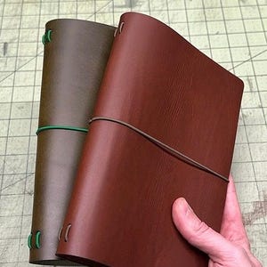 May include: Two rectangular leather journals, one in a dark brown shade and the other in a lighter reddish-brown tone. Each journal is secured with a thin elastic band wrapped horizontally around the cover and features visible elastic binding loops.