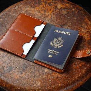 May include: A tan leather passport holder with visible stitching, featuring two interior card slots on the left side and a dark blue United States of America passport inserted into the right side, secured by a small metal snap button closure.