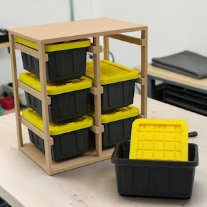 May include: A wooden storage shelf unit with six black bins with yellow lids. One bin is separate, with its lid off. The unit is on a light-colored surface, suggesting a workshop or storage space.