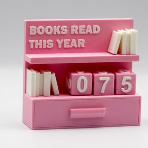 Puede incluir: Un organizador de escritorio rosa con forma de estanter&iacute;a con el texto "BOOKS READ THIS YEAR". Tiene dos estantes con pilas de libros en miniatura y bloques numerados. El caj&oacute;n muestra el n&uacute;mero 075.