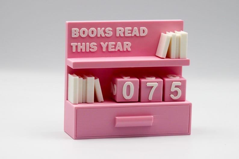 Puede incluir: Un organizador de escritorio rosa con forma de estanter&iacute;a con el texto "BOOKS READ THIS YEAR". Tiene dos estantes con pilas de libros en miniatura y bloques numerados. El caj&oacute;n muestra el n&uacute;mero 075.