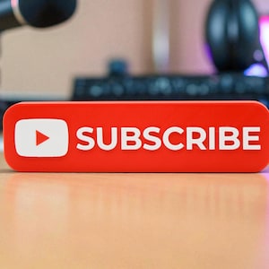 May include: A red rectangular sign with rounded corners, featuring the YouTube logo and the word "SUBSCRIBE" in white capital letters. The sign is on a wooden surface, with a microphone and other tech equipment in the background.