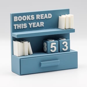 Puede incluir: Un organizador de escritorio azul con forma de estanter&iacute;a, con el texto "BOOKS READ THIS YEAR" en blanco. Cuenta con pilas de libros en miniatura en estantes y bloques de n&uacute;meros que muestran "853". Un caj&oacute;n est&aacute; en la parte inferior.