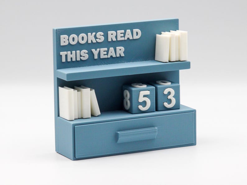 Puede incluir: Un organizador de escritorio azul con forma de estanter&iacute;a, con el texto "BOOKS READ THIS YEAR" en blanco. Cuenta con pilas de libros en miniatura en estantes y bloques de n&uacute;meros que muestran "853". Un caj&oacute;n est&aacute; en la parte inferior.
