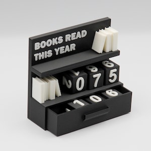 Puede incluir: Un organizador de escritorio negro con forma de estanter&iacute;a, con el texto "BOOKS READ THIS YEAR" en blanco. Cuenta con peque&ntilde;as pilas de libros blancos y bloques num&eacute;ricos para rastrear el progreso de lectura. Los n&uacute;meros son 0, 7, 5, 1, 0 y 6.