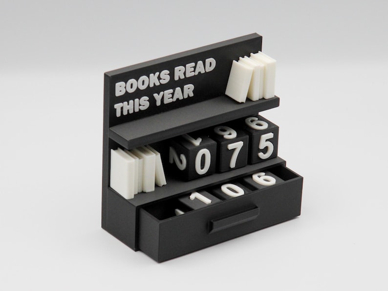 Puede incluir: Un organizador de escritorio negro con forma de estanter&iacute;a, con el texto "BOOKS READ THIS YEAR" en blanco. Cuenta con peque&ntilde;as pilas de libros blancos y bloques num&eacute;ricos para rastrear el progreso de lectura. Los n&uacute;meros son 0, 7, 5, 1, 0 y 6.
