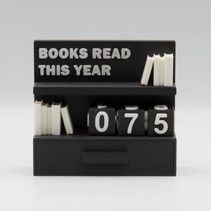 Puede incluir: Un mostrador negro con forma de estanter&iacute;a con el texto "BOOKS READ THIS YEAR" en blanco. Tiene dos estantes con peque&ntilde;as pilas de libros blancos y un contador que muestra "075". Hay un caj&oacute;n en la parte inferior.