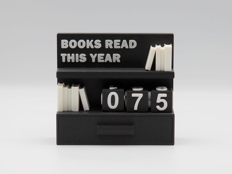 Puede incluir: Un mostrador negro con forma de estanter&iacute;a con el texto "BOOKS READ THIS YEAR" en blanco. Tiene dos estantes con peque&ntilde;as pilas de libros blancos y un contador que muestra "075". Hay un caj&oacute;n en la parte inferior.
