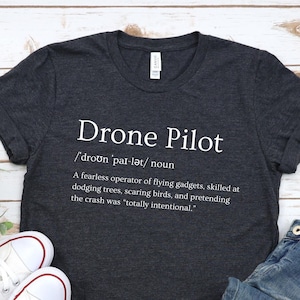 May include: A dark gray t-shirt with the text "Drone Pilot" and a definition of the term. The shirt is laid flat on a white wooden surface with a pair of white canvas shoes and blue jeans.