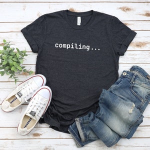 May include: A dark grey t-shirt with the text "compiling..." in white on the front. The shirt is laid out on a white wooden surface with a pair of white trainers and blue jeans.