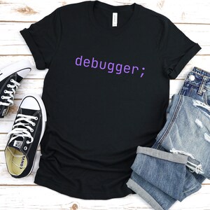 Könnte beinhalten: Ein schwarzes T-Shirt mit dem Text "debugger;" in lila auf der Vorderseite. Das Shirt liegt flach auf einer weißen Holzoberfläche mit einem Paar schwarzen Turnschuhen und einer blauen Jeans.