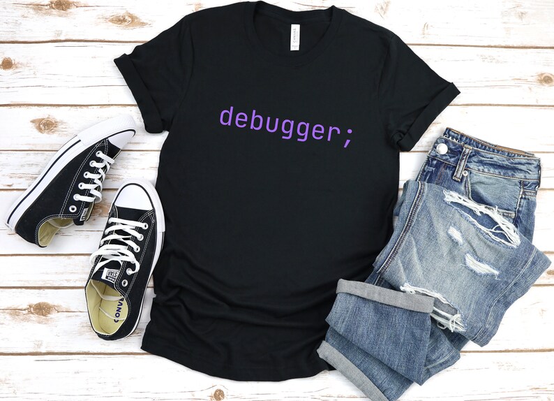 May include: A black t-shirt with the text "debugger;" printed in purple on the front. The shirt is laid flat on a white wooden surface with a pair of black trainers and a pair of blue jeans.