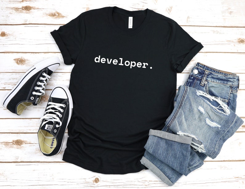 Peut inclure: T-shirt noir avec le texte "developer." en lettres blanches. Le t-shirt est pos&eacute; &agrave; plat sur une surface en bois blanc avec une paire de baskets noires et un jean bleu.