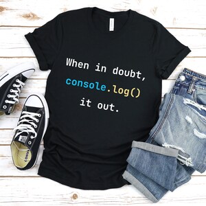 May include: Black t-shirt with the text "When in doubt, console.log() it out." in white, blue, and yellow. The shirt is laid flat on a white wooden surface with a pair of black Converse shoes and blue jeans.