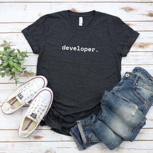 Peut inclure: Un t-shirt gris fonc&eacute; avec le mot "developer." imprim&eacute; en blanc sur le devant. Le t-shirt est pos&eacute; &agrave; plat sur une surface en bois blanc avec une paire de chaussures en toile blanche et un jean bleu.