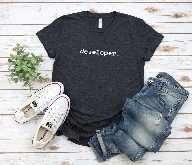Peut inclure: Un t-shirt gris fonc&eacute; avec le mot "developer." imprim&eacute; en blanc sur le devant. Le t-shirt est pos&eacute; &agrave; plat sur une surface en bois blanc avec une paire de chaussures en toile blanche et un jean bleu.
