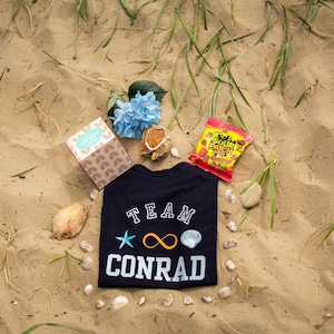 May include: A navy blue t-shirt with the text "TEAM CONRAD" in white, featuring a starfish, infinity symbol, and seashell. The shirt is on sand, with seashells, a box, sweets, and blue flowers scattered around.