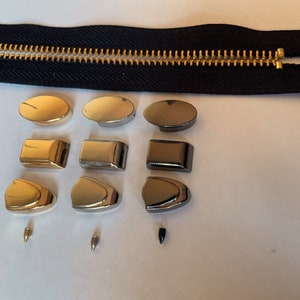 May include: Assortment of zipper pulls in gold, silver, and black metal finishes. The pulls are in various shapes, including oval, rectangular, and shield-shaped. Some pulls have a screw-on design.