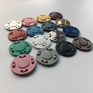 May include: Assortment of 14 round metal snap buttons in various colours, including red, green, blue, brown, gold, silver, and white. The buttons have a raised centre and a ring of small holes around the edge.