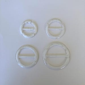 May include: Four white resin buckles with a marbled effect. The buckles are oval and circular, with a bar across the centre. The sizes vary, with two smaller and two larger buckles. The buckles are on a white background.