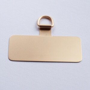 May include: A gold-colored metal tag with a rectangular shape and a small loop at the top. The tag is blank.