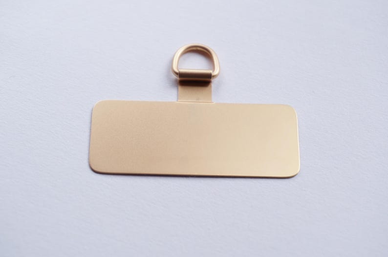 May include: A gold-colored metal tag with a rectangular shape and a small loop at the top. The tag is blank.