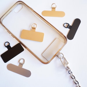 May include: A clear phone case with a gold and silver chain attached. The case has a gold and black rectangular tag attached to it. There are three additional tags, one gold and two black, on a white surface.