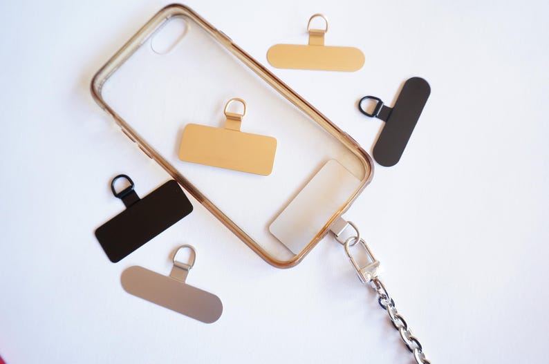 May include: A clear phone case with a gold and silver chain attached. The case has a gold and black rectangular tag attached to it. There are three additional tags, one gold and two black, on a white surface.