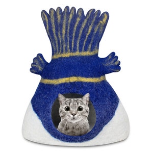 以下が含まれることがあります： 黄色い縁取りのある帽子型の青と白のフェルト製の猫用ベッド。グレーのトラ猫がベッドの開口部から外を覗いています。