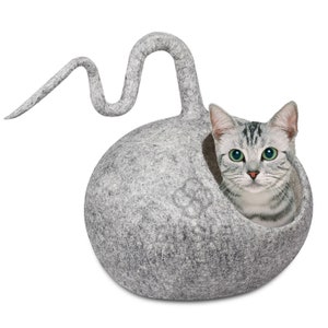 以下が含まれることがあります： 長い尻尾のある丸まった猫の形をした灰色のフェルト製の猫用ベッド。グレーと白の虎柄の猫がベッドから顔を出しています。