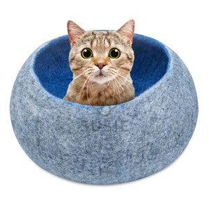 以下が含まれることがあります： 青いフェルト製の猫用ベッドから、緑色の目をした茶トラ猫が顔をのぞかせている。