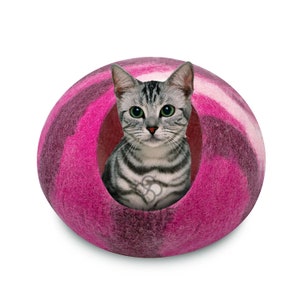 以下が含まれることがあります： グレーと白の虎柄の猫が、ピンクと白のフェルト製の猫用ベッドの中に座っています。ベッドは球形で、猫が出入りできる小さな開口部があります。